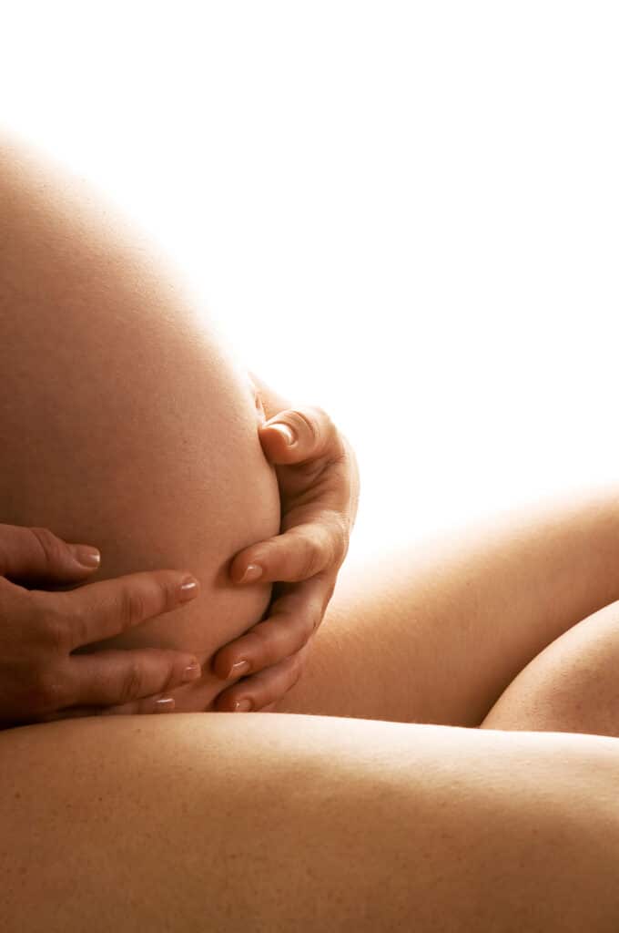 Mains tenant ventre grossesse peau douce fond lumineux Gros plan d'une femme enceinte tenant son ventre arrondi de ses mains, peau bronzée sur fond blanc lumineux.